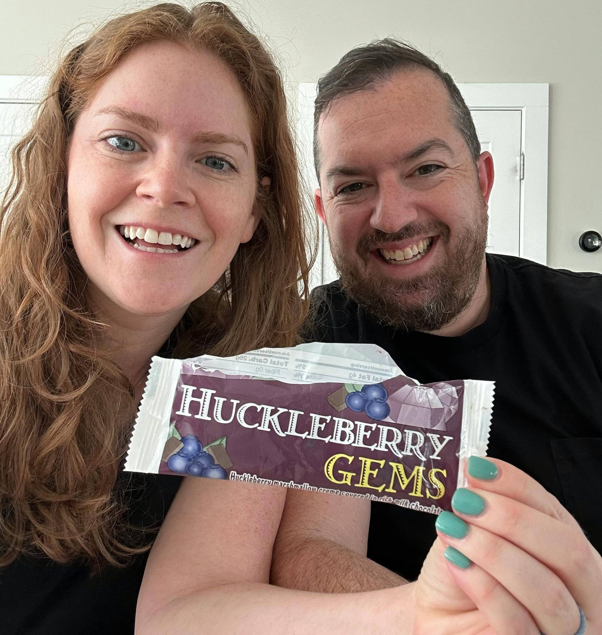 A couple smiling at the camera, holding a candy wrapper up that says "Huckleberry Gems"